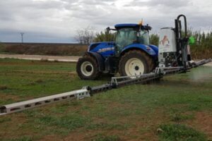 Tractor agrícola azul New Holland con pulverizador de barras extendidas aplicando fitosanitarios en un campo, bajo un cielo nublado, simbolizando una aplicación cualificada.