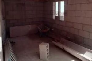 Un espacio interior en construcción con paredes de bloques de cemento, un techo con vigas de madera expuestas y un suelo de hormigón. Se ven elementos de obra como una carretilla y una escalera apoyada en una pared. Una ventana rectangular en la pared del fondo permite la entrada de luz.
