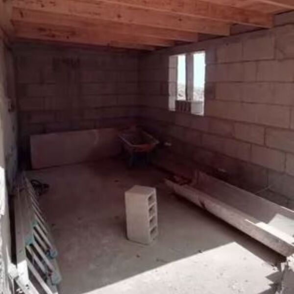 Un espacio interior en construcción con paredes de bloques de cemento, un techo con vigas de madera expuestas y un suelo de hormigón. Se ven elementos de obra como una carretilla y una escalera apoyada en una pared. Una ventana rectangular en la pared del fondo permite la entrada de luz.