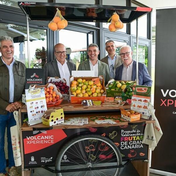 Cinco hombres sonrientes posan alrededor de un atractivo stand de mercado lleno de frutas frescas como naranjas, limones y uvas, con el logo 